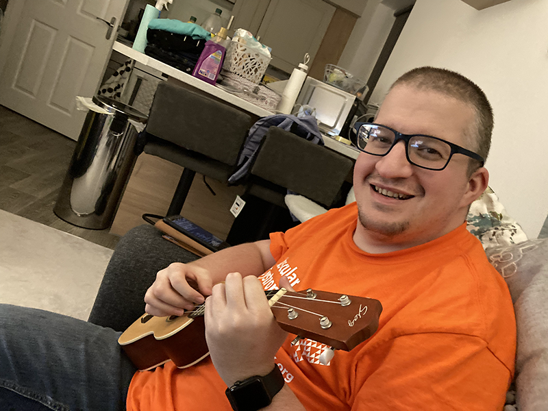 me sat on sofa, orange t-shirt, playing ukulele